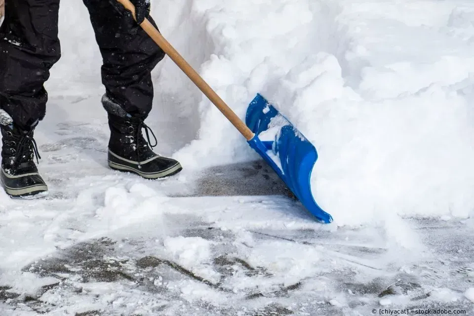 Eine Person schaufelt mit einer blauen Schneeschaufel Schnee auf einer verschneiten Einfahrt.