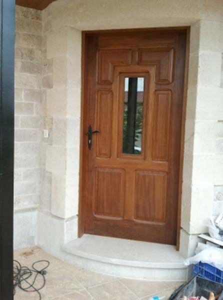 Puerta de entrada de madera con panel de vidrio, marco de piedra color tostado, colocada en una pared de piedra beige.