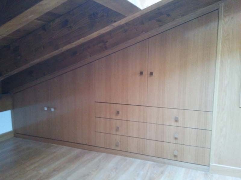 Muebles empotrados de madera bajo techo inclinado; cajones y puertas de armarios, de madera clara.