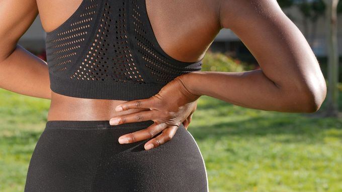 Persona con ropa deportiva negra, conteniéndose, probablemente con dolor. Al aire libre, en un entorno soleado.