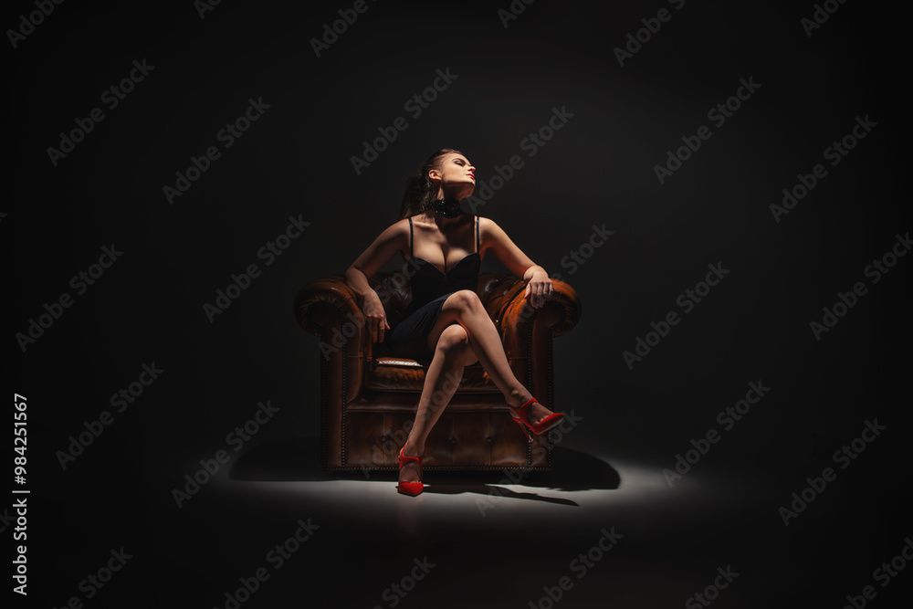 Eine Person in einem schwarzen Kleid sitzt in einem Ledersessel unter einem Scheinwerfer vor einem dunklen Hintergrund.