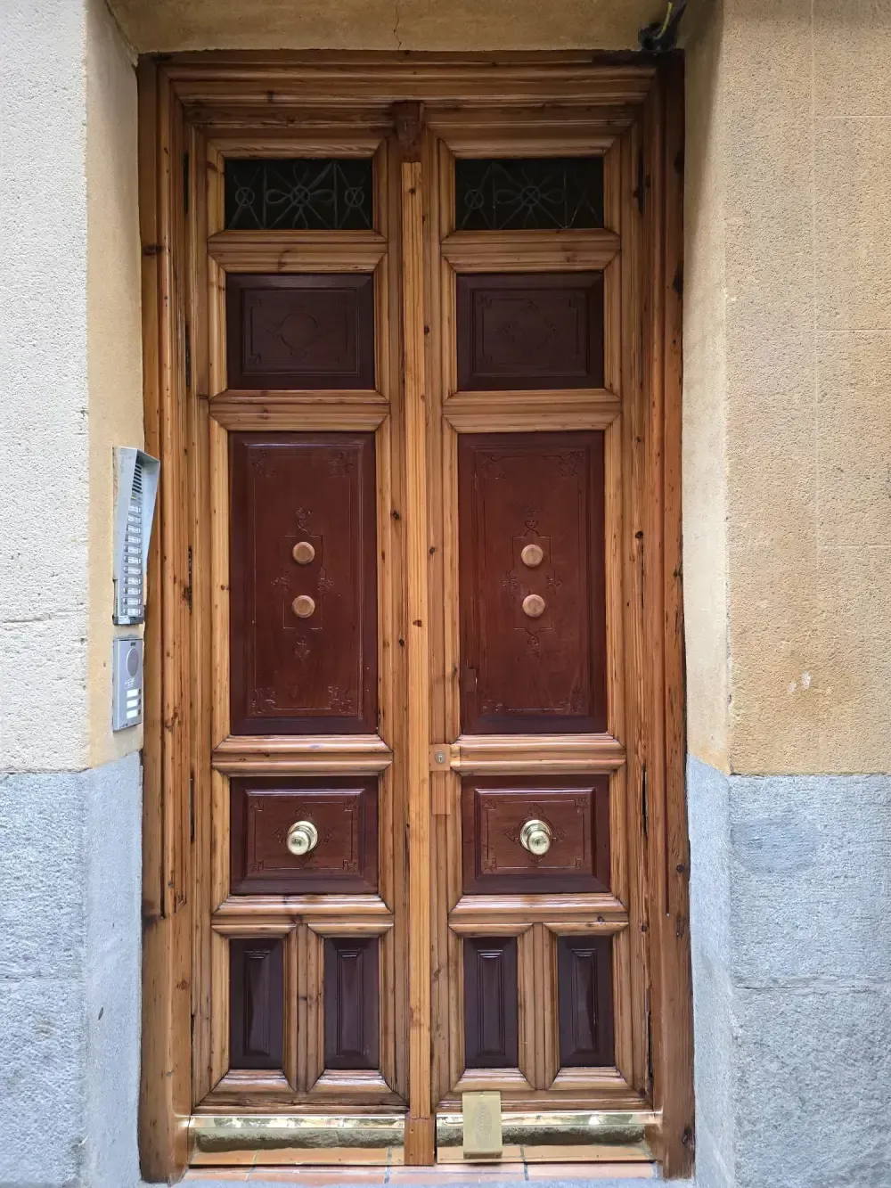Puertas dobles de madera con paneles de color marrón oscuro, herrajes dorados y una pared de piedra y beige.