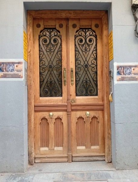 Puertas dobles de madera con herrajes ornamentados, colocadas en una pared gris claro.