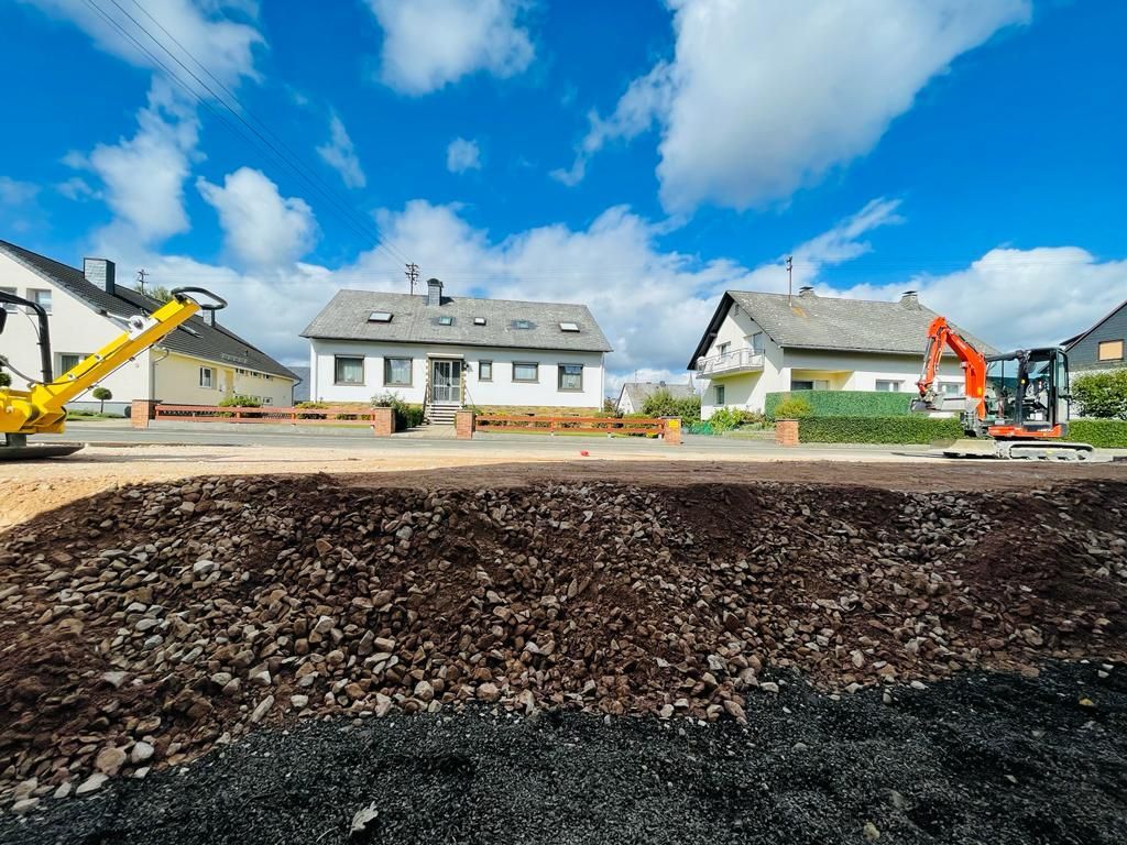 Eine Baustelle mit einem Haus im Hintergrund und einem gelben Bagger im Vordergrund.