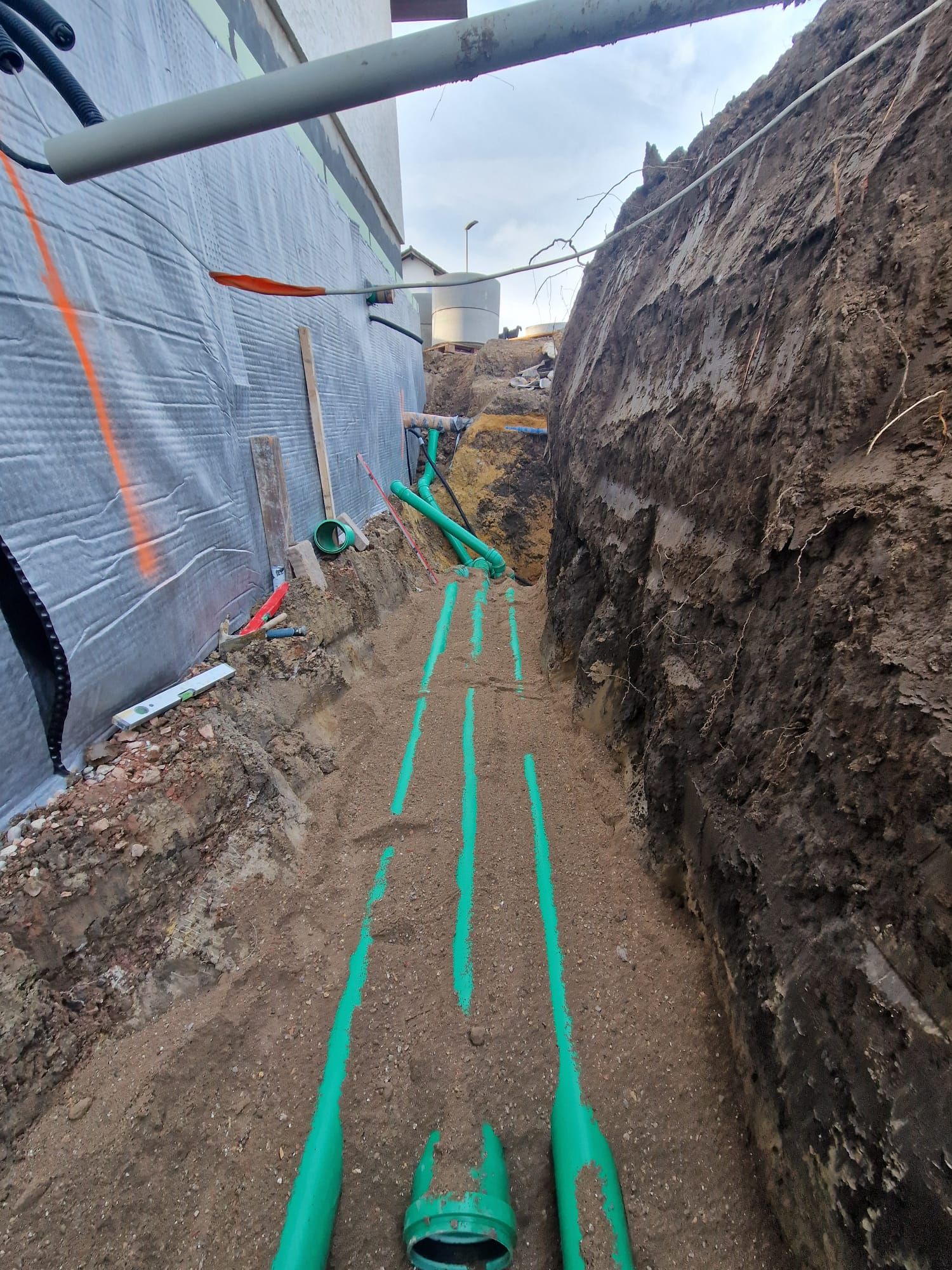 In einem Graben neben einem Gebäude werden grüne Rohre verlegt.