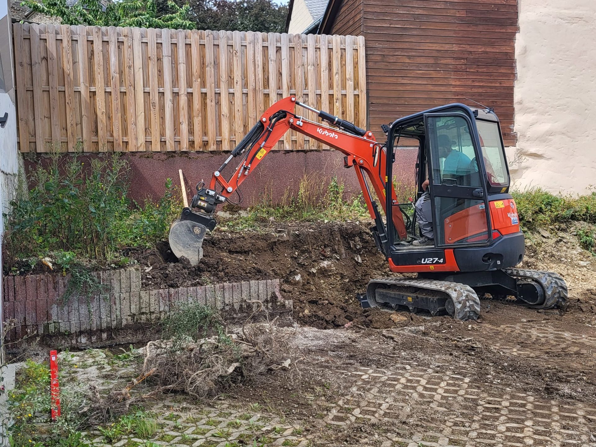 Ein kleiner Bagger gräbt vor einem Holzzaun ein Loch in die Erde.
