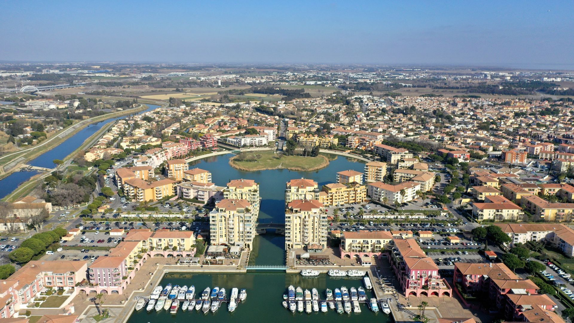 Vue aérienne d'un quartier riverain d'un canal avec des bâtiments, des bateaux et un port de plaisance sous un ciel dégagé.