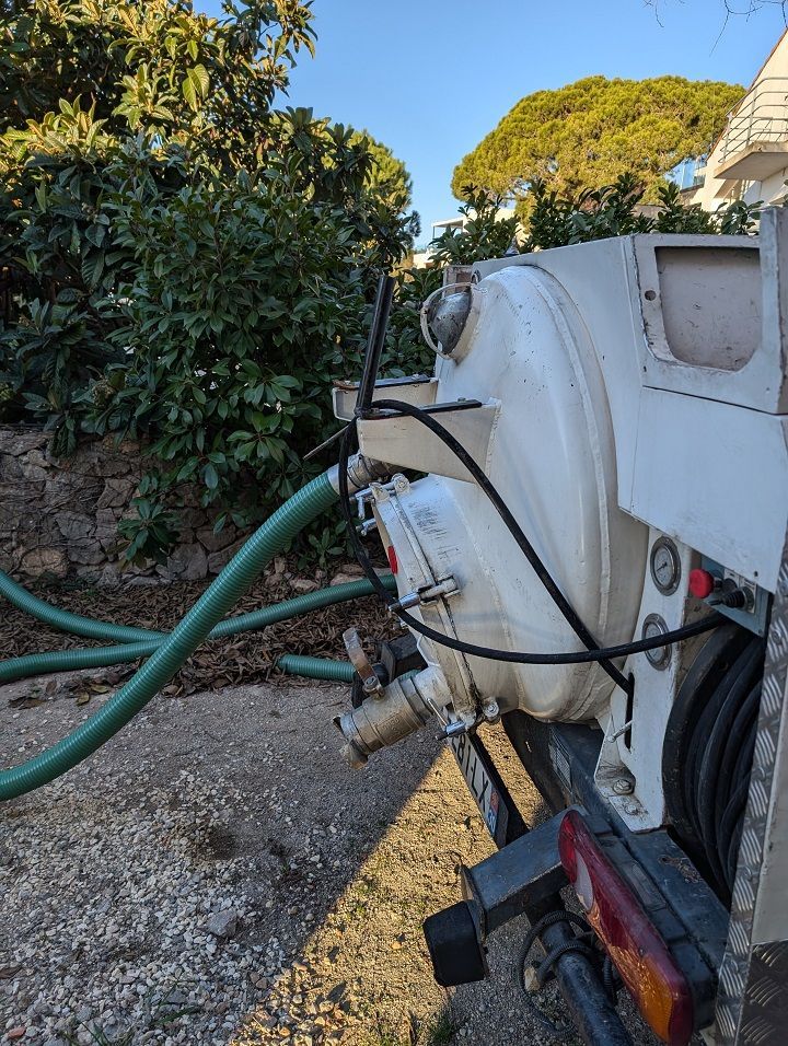 Réservoir de pompe blanc avec tuyau vert dans une cour gravillonnée, à côté de buissons denses et d'une maison