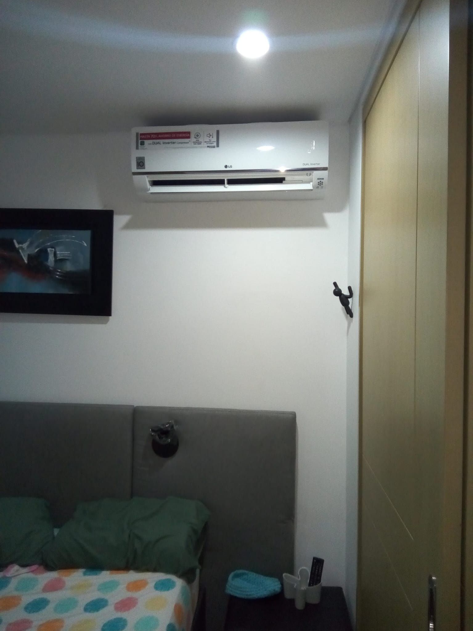 Habitación pequeña con cama, televisor montado en la pared y aire acondicionado encima del cabecero.