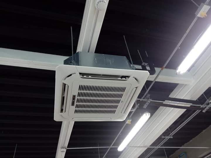 Unidad de aire acondicionado blanca montada en el techo en un espacio interior industrial con luces fluorescentes.