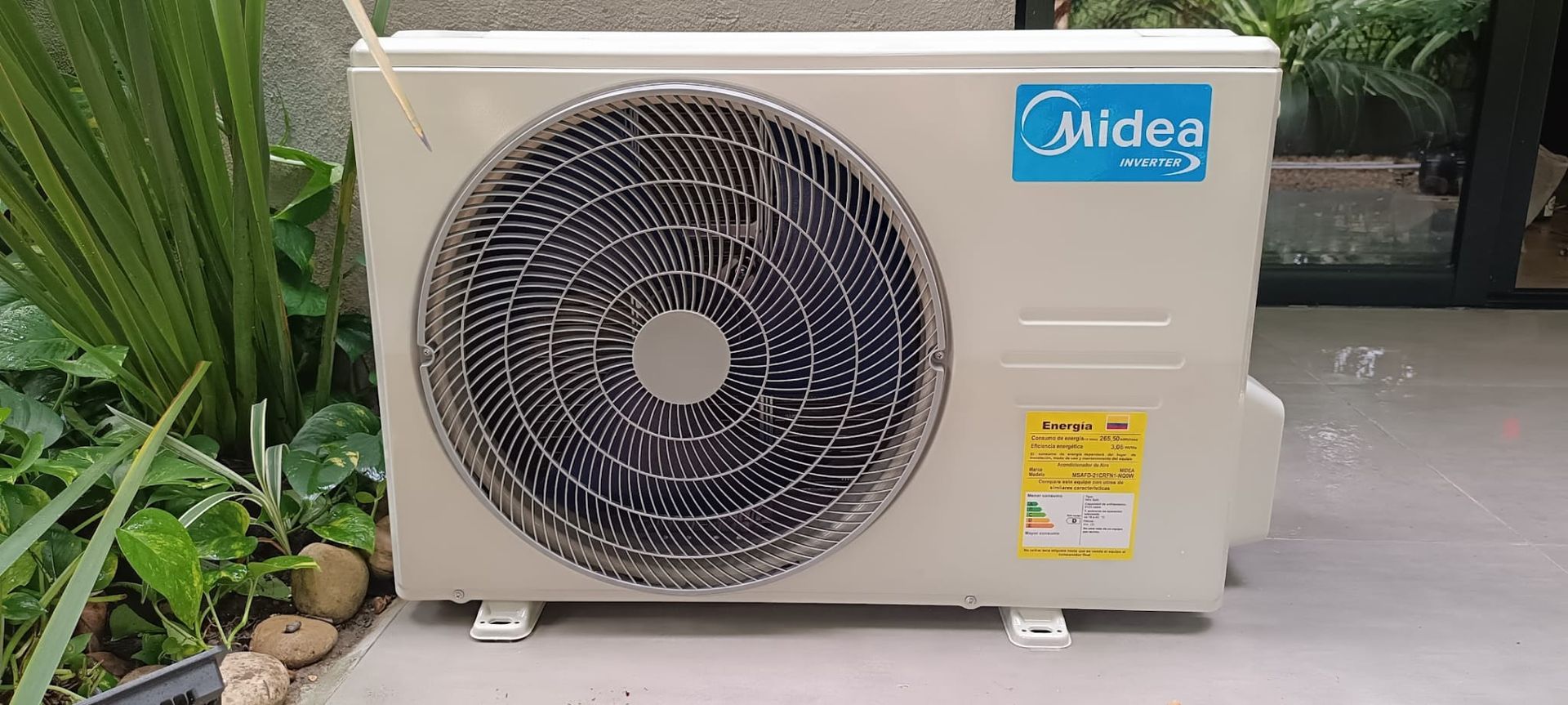 Unidad de aire acondicionado exterior Midea junto a plantas en un patio.