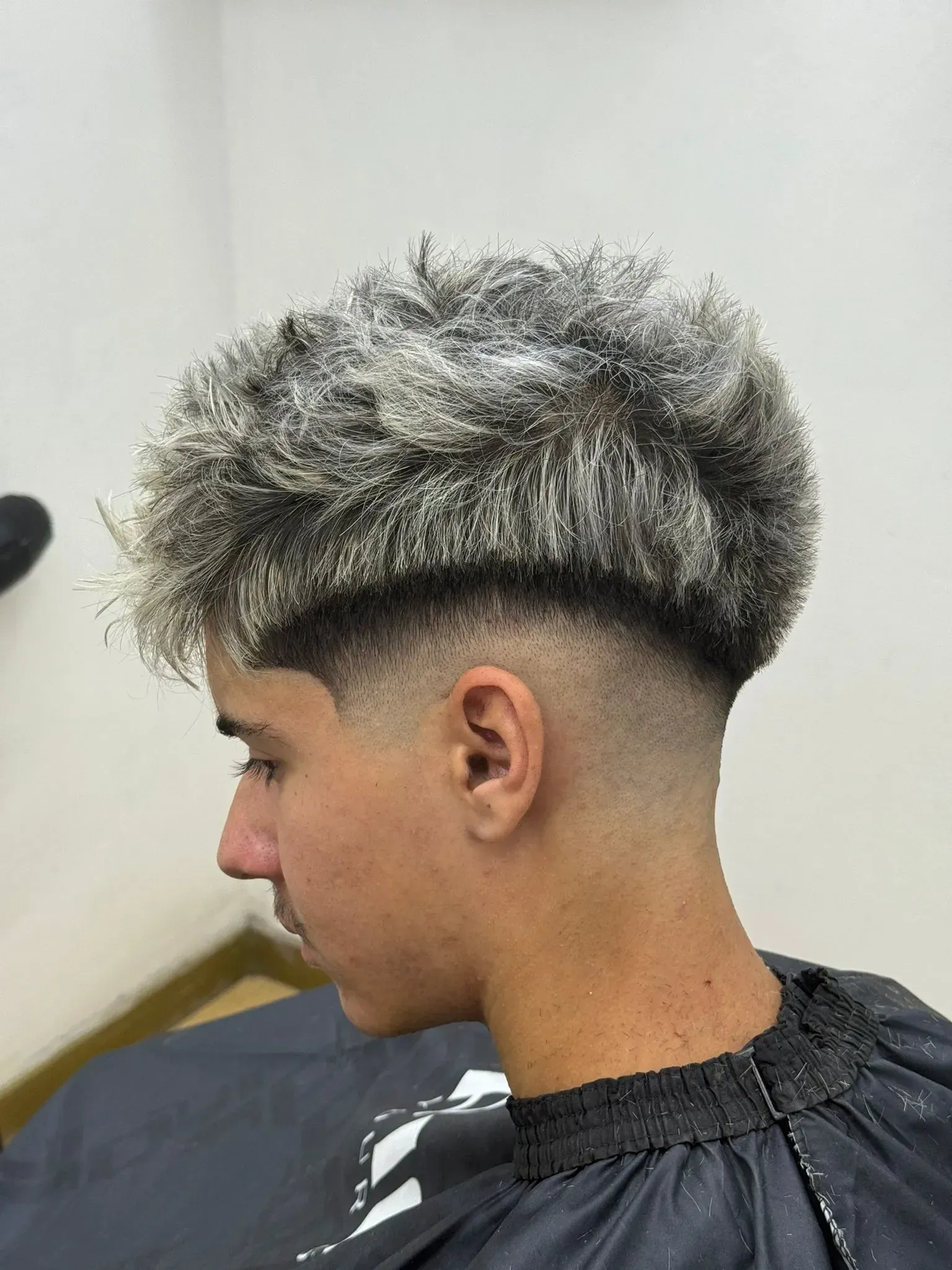 Adolescente con cabello texturizado de color claro, corte desvanecido, perfil lateral, vistiendo una capa de salón.