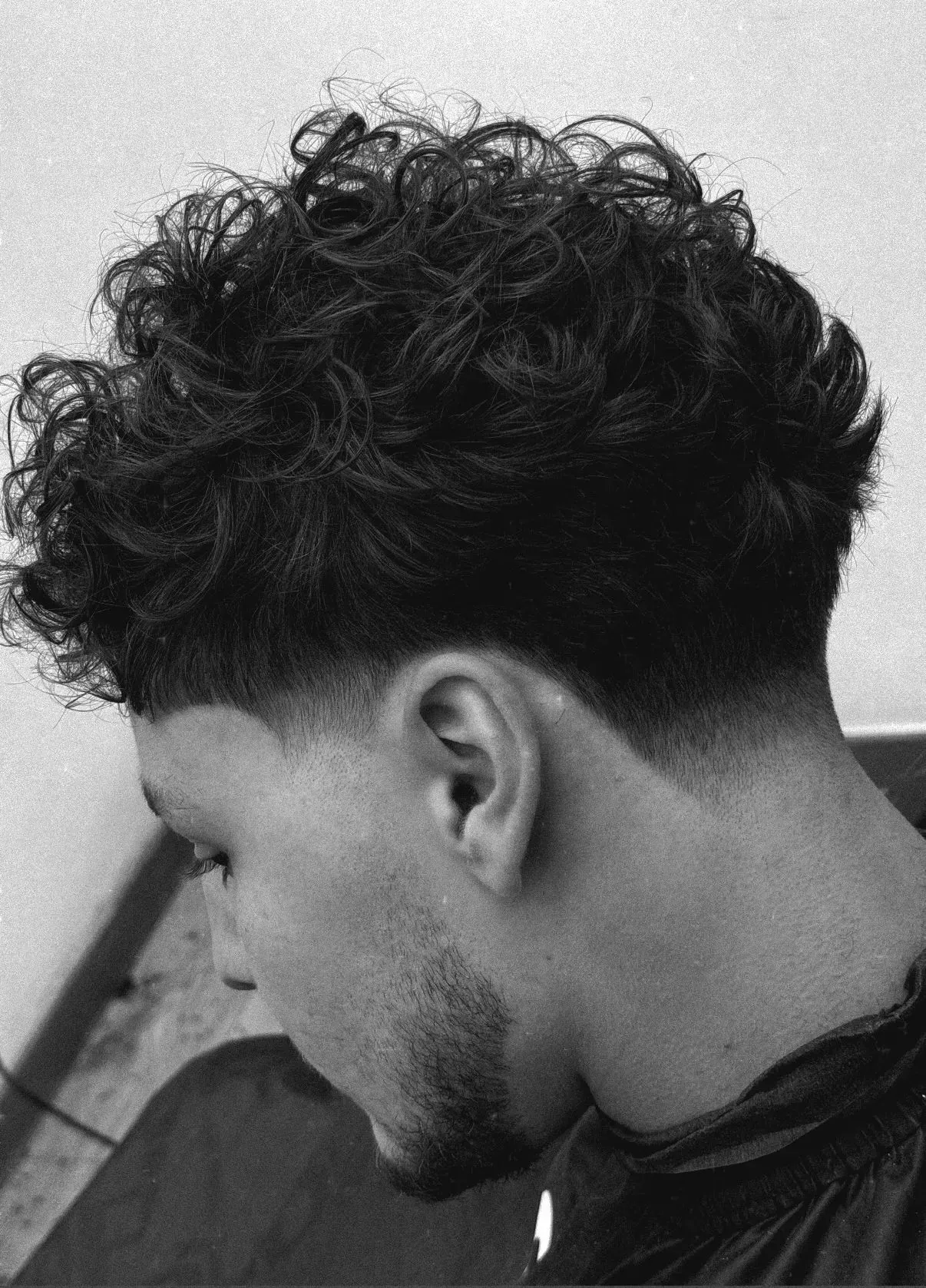 Hombre con cabello rizado y corte desvanecido, mirando hacia abajo. Blanco y negro.