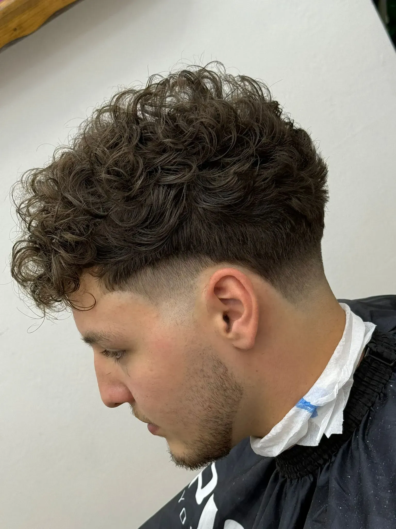 Hombre con cabello rizado y corte de pelo descolorido, mirando hacia abajo.