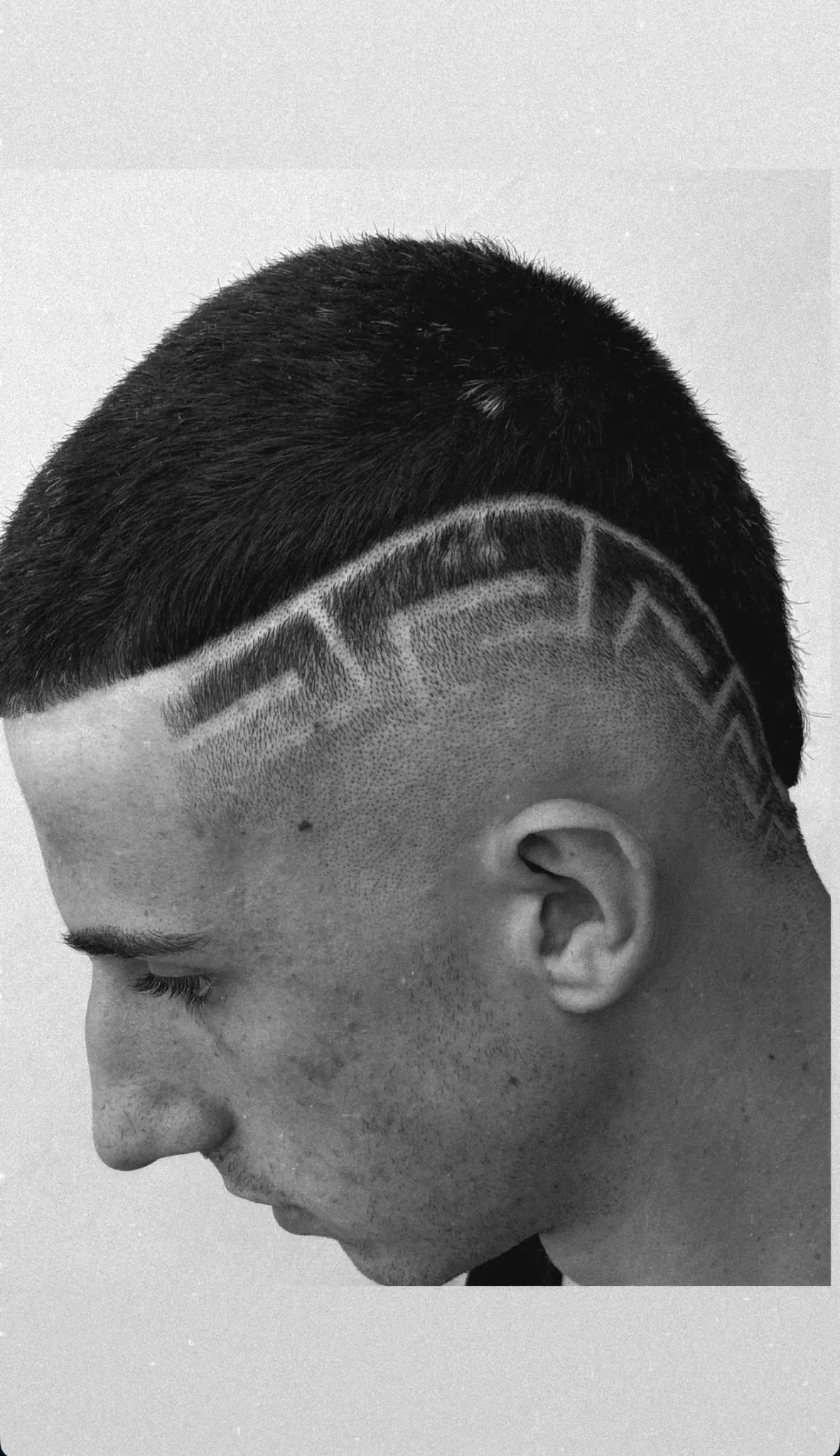 Hombre con la cabeza rapada y un diseño estampado en el costado de la cabeza, mirando hacia abajo.