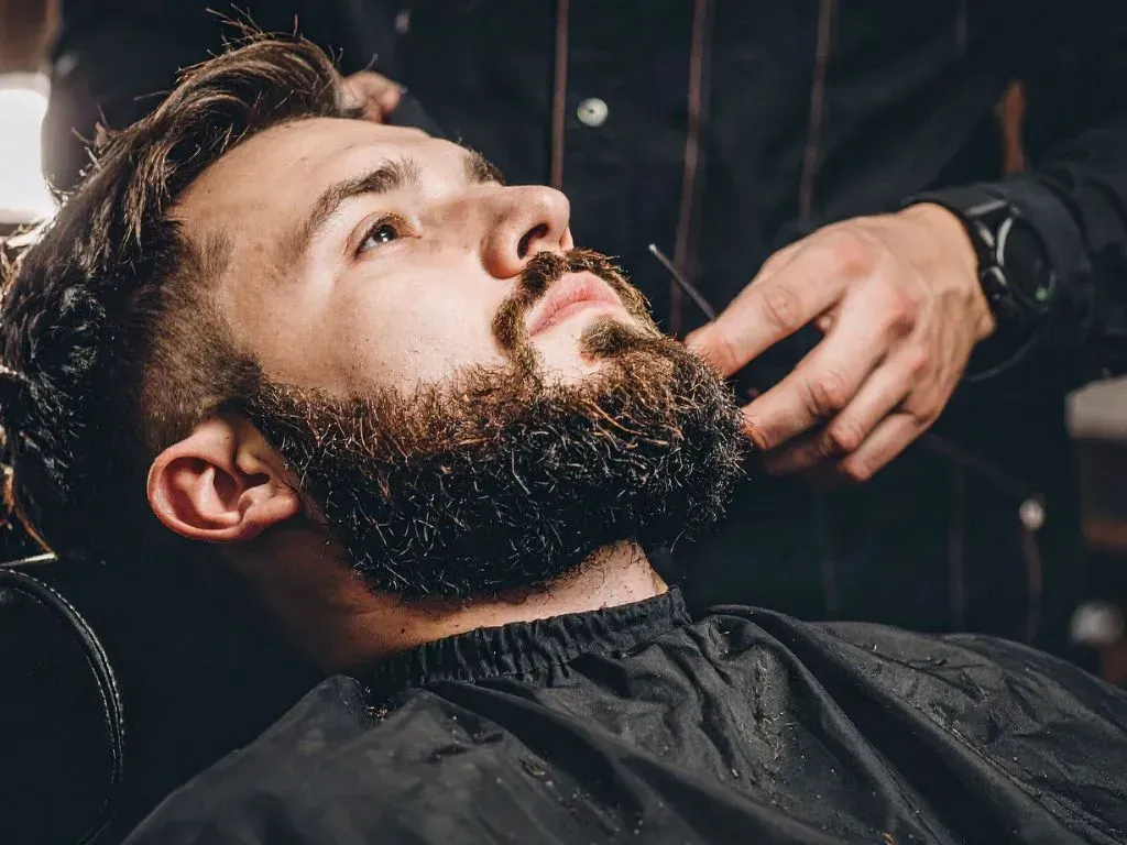 Un barbero le corta la barba a un hombre con tijeras. El hombre está reclinado en una silla de peluquería.
