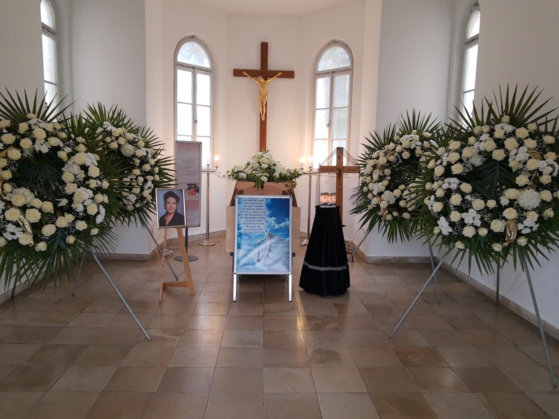 Sarg umgeben von großen Blumenarrangements und Kreuz.