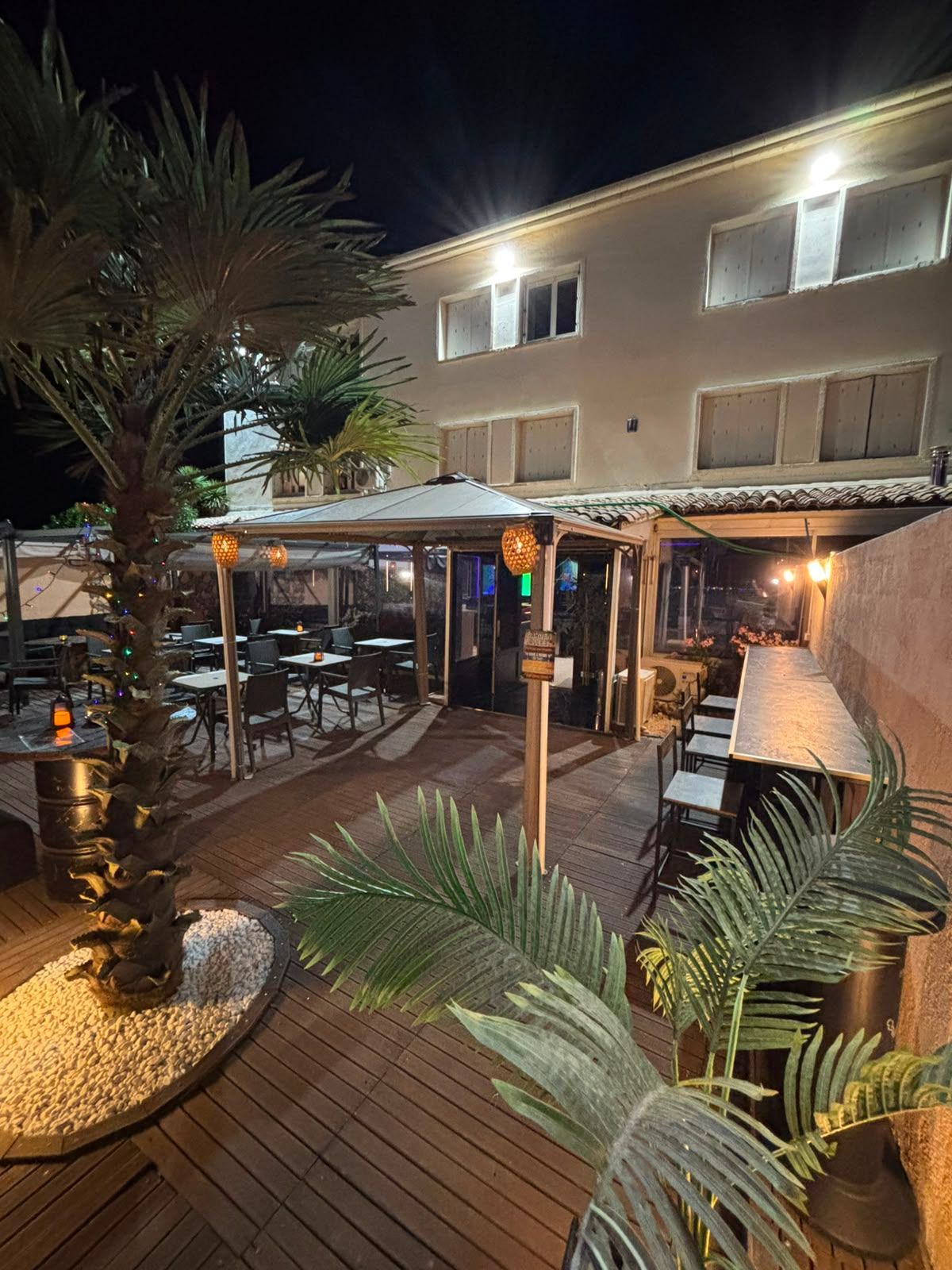 Terrasse de restaurant extérieure nocturne avec plancher en bois, palmiers, tables et guirlandes lumineuses chaleureuses près d'un bâtiment