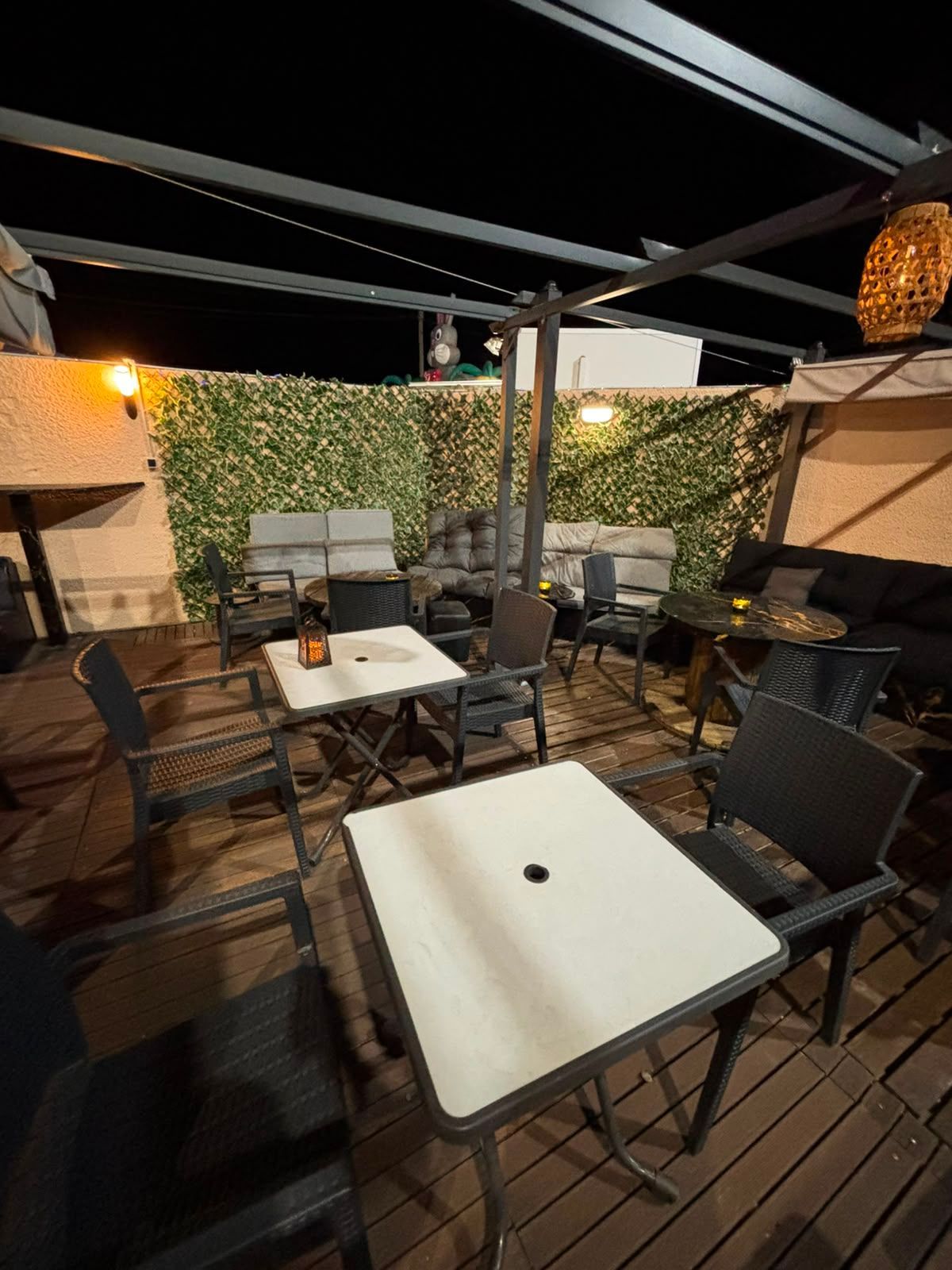 Terrasse de café vide avec chaises noires, tables blanches et haie éclairée par des guirlandes lumineuses.