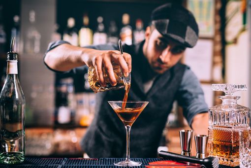 Un barman verse un cocktail dans un verre à martini derrière un bar rempli de bouteilles d'alcool.