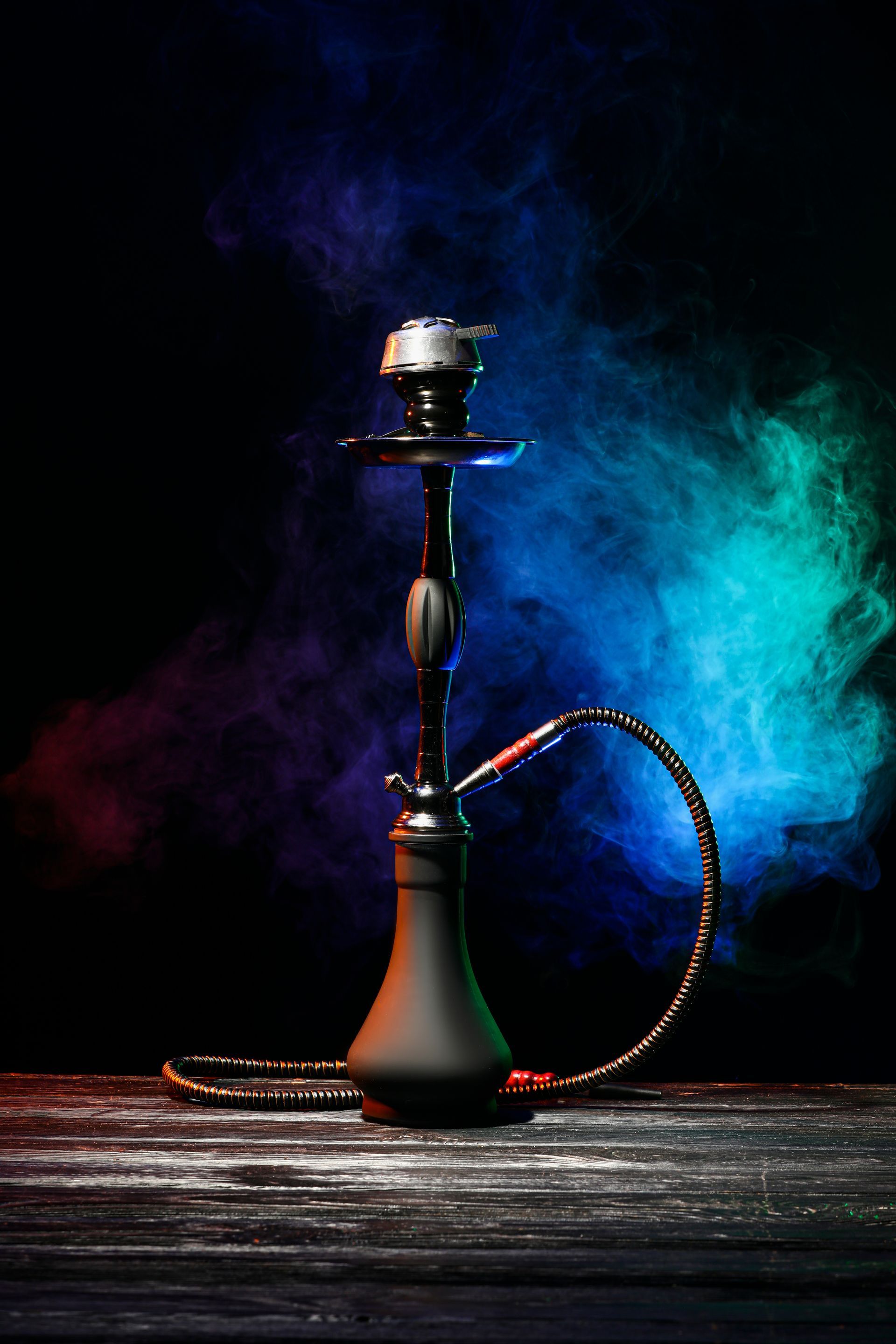 Narguilé à tige noire et base en verre posé sur une table en bois, sur fond de fumée bleue et violette.