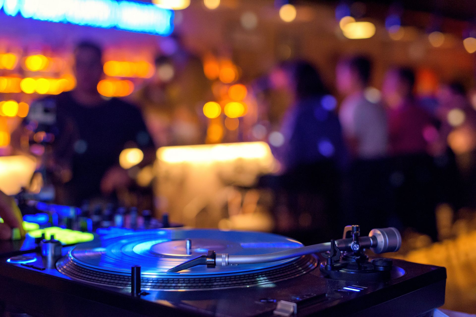 Platine de DJ avec des lumières bleues scintillantes dans une boîte de nuit, avec des silhouettes floues et des lumières chaudes en arrière-plan.