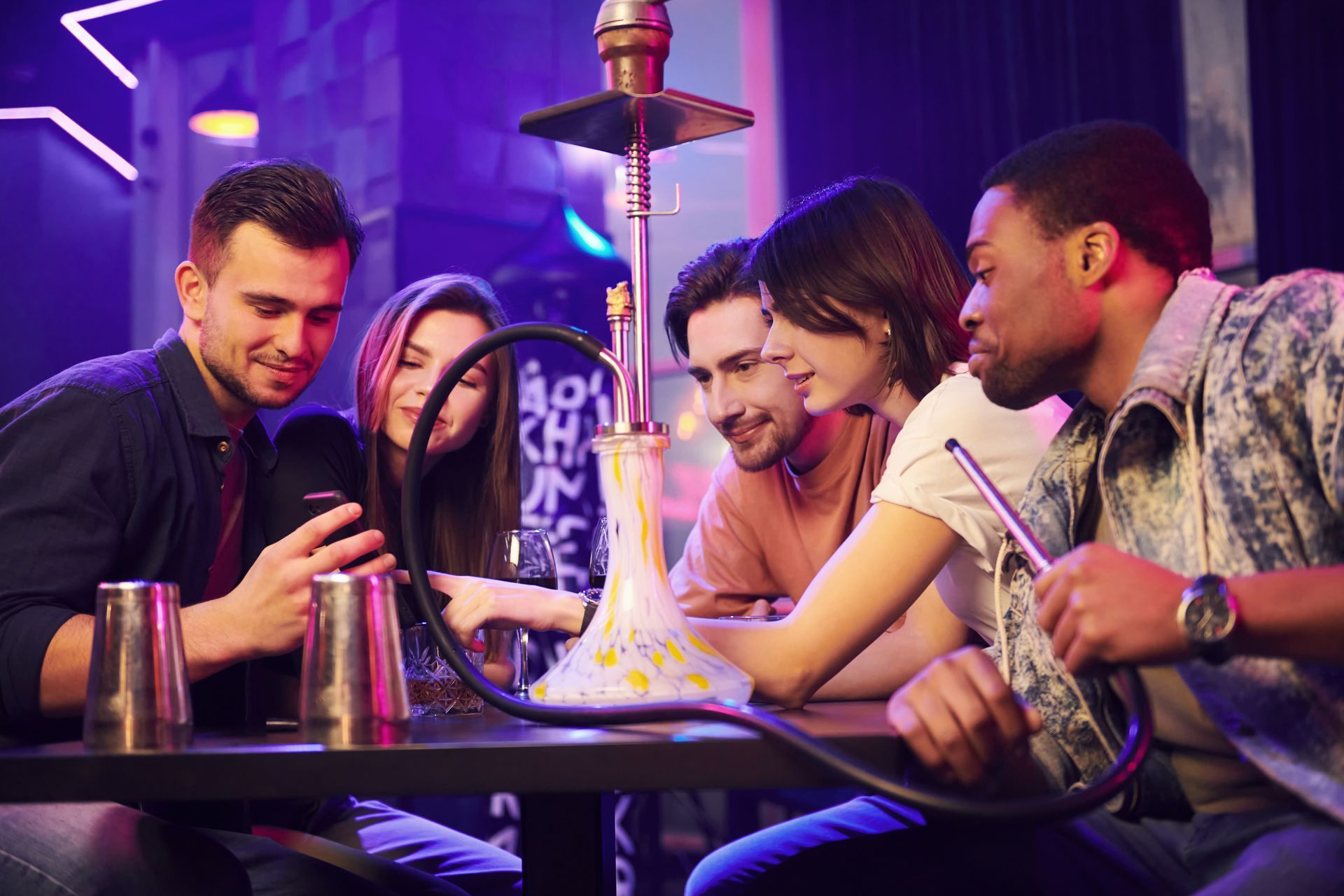 Quatre amis discutent autour d'une table à narguilé dans un salon à l'éclairage tamisé et violet.