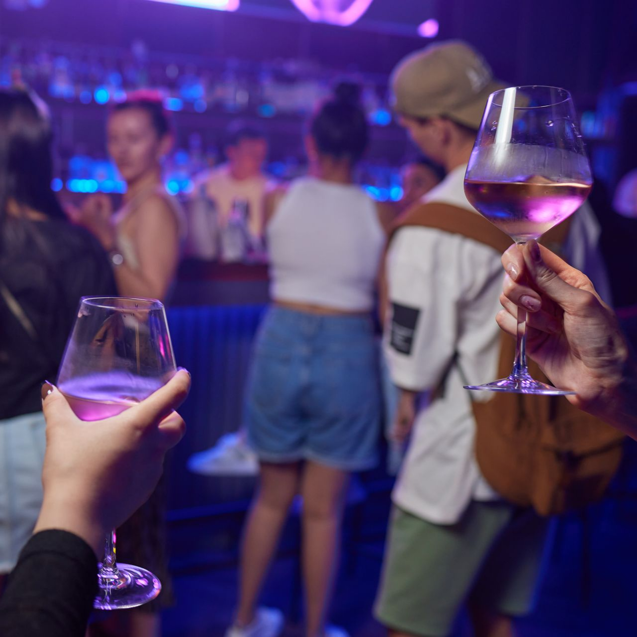 Des mains tenant des verres à vin dans un bar éclairé au néon, avec des gens qui discutent en arrière-plan flou.