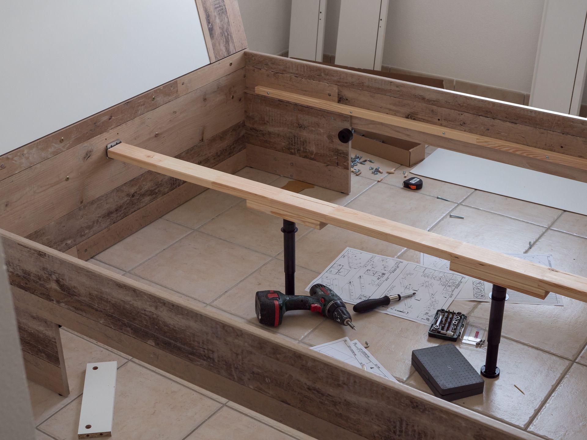 Cadre de lit en bois partiellement assemblé, outils, instructions, sur sol carrelé.