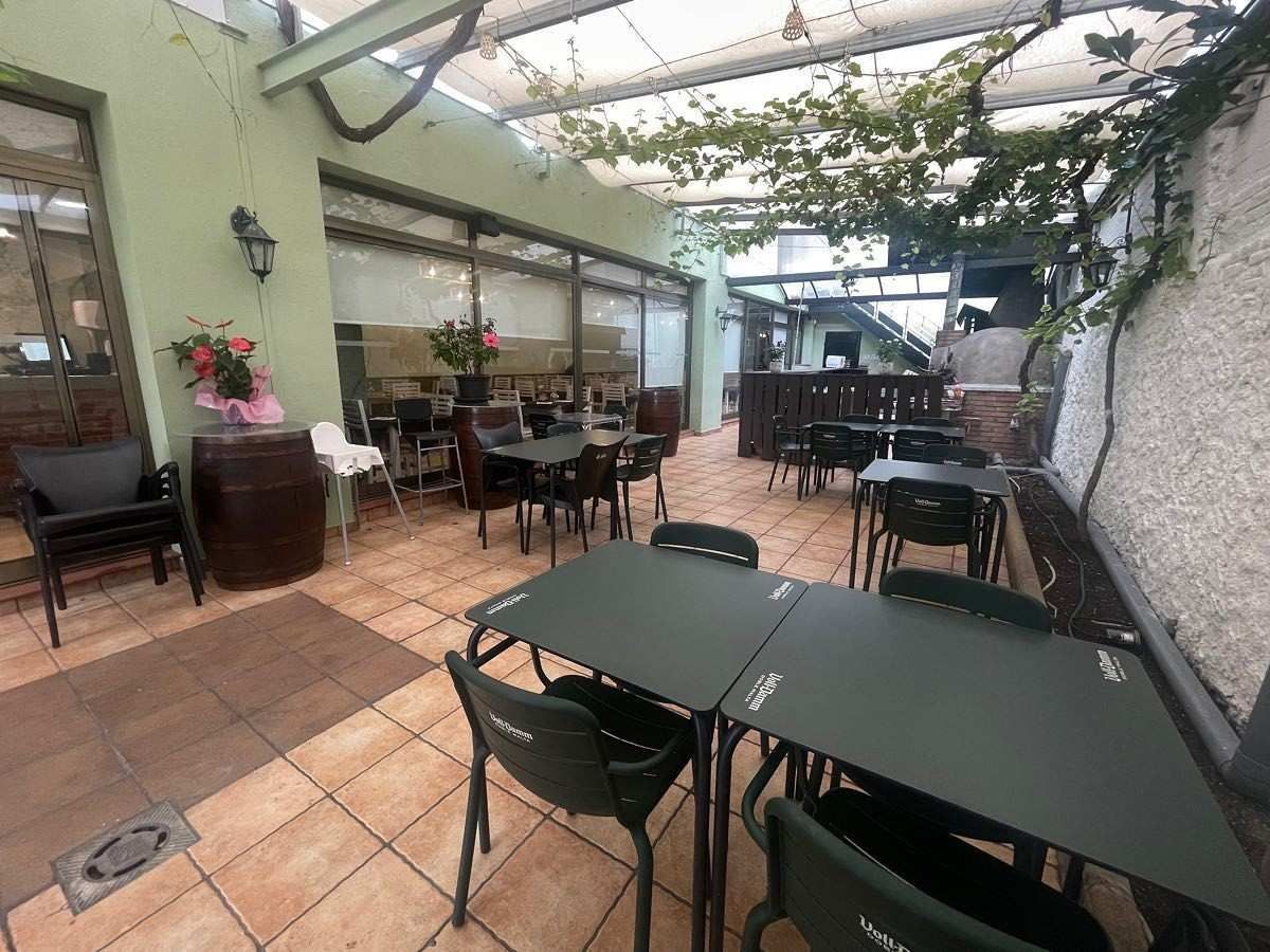 Terraza de cafetería al aire libre con mesas y sillas verdes bajo un toldo cubierto de enredaderas.