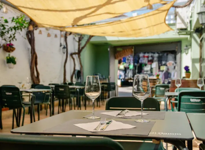 Terraza de cafetería bajo un toldo, con mesas dispuestas y copas de vino en un patio soleado.