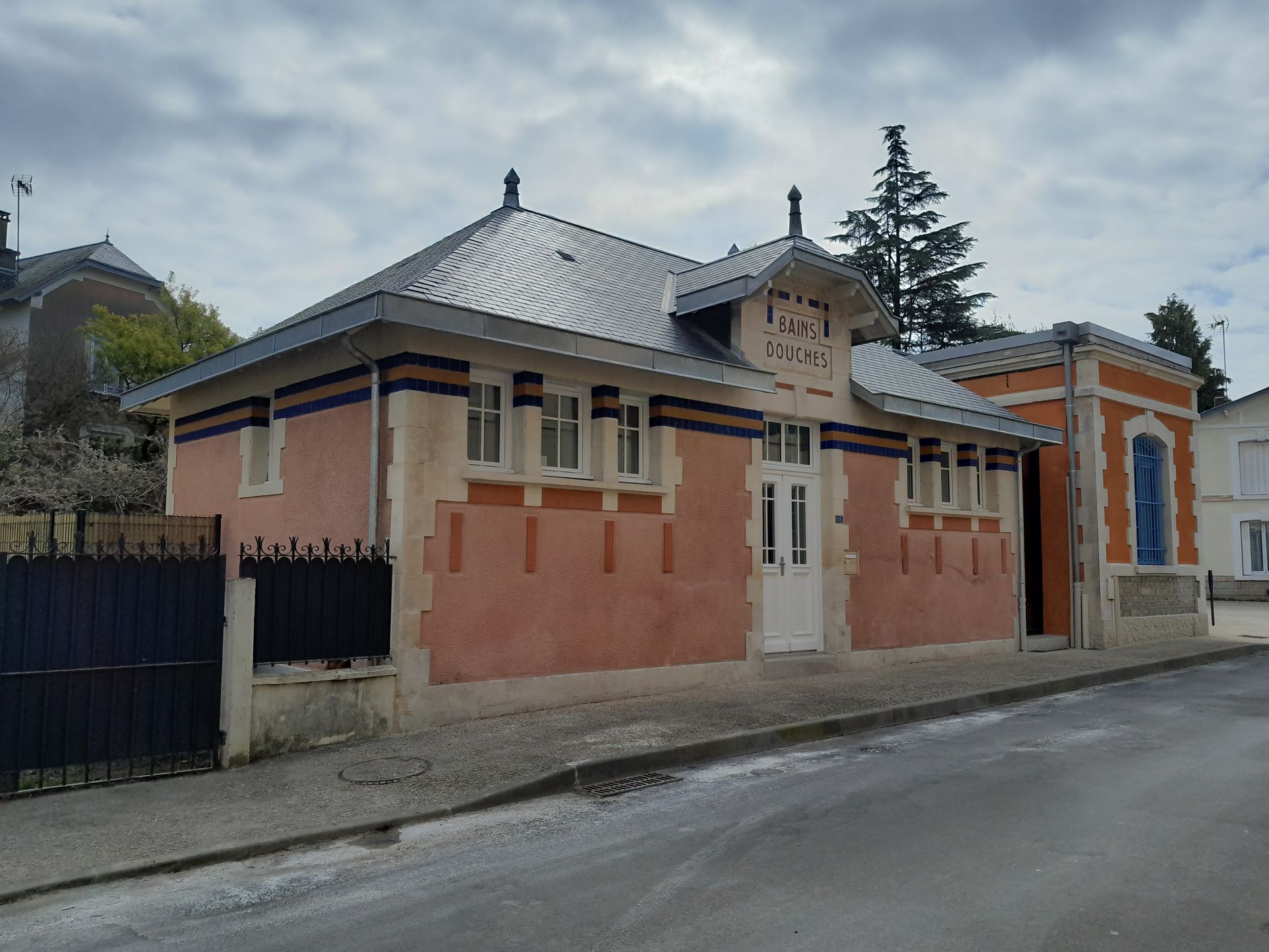 Vue depuis la rue d’une maison aux tons pêche et gris, avec un toit en métal et une entrée fermée par un portail, sur une route tranquille