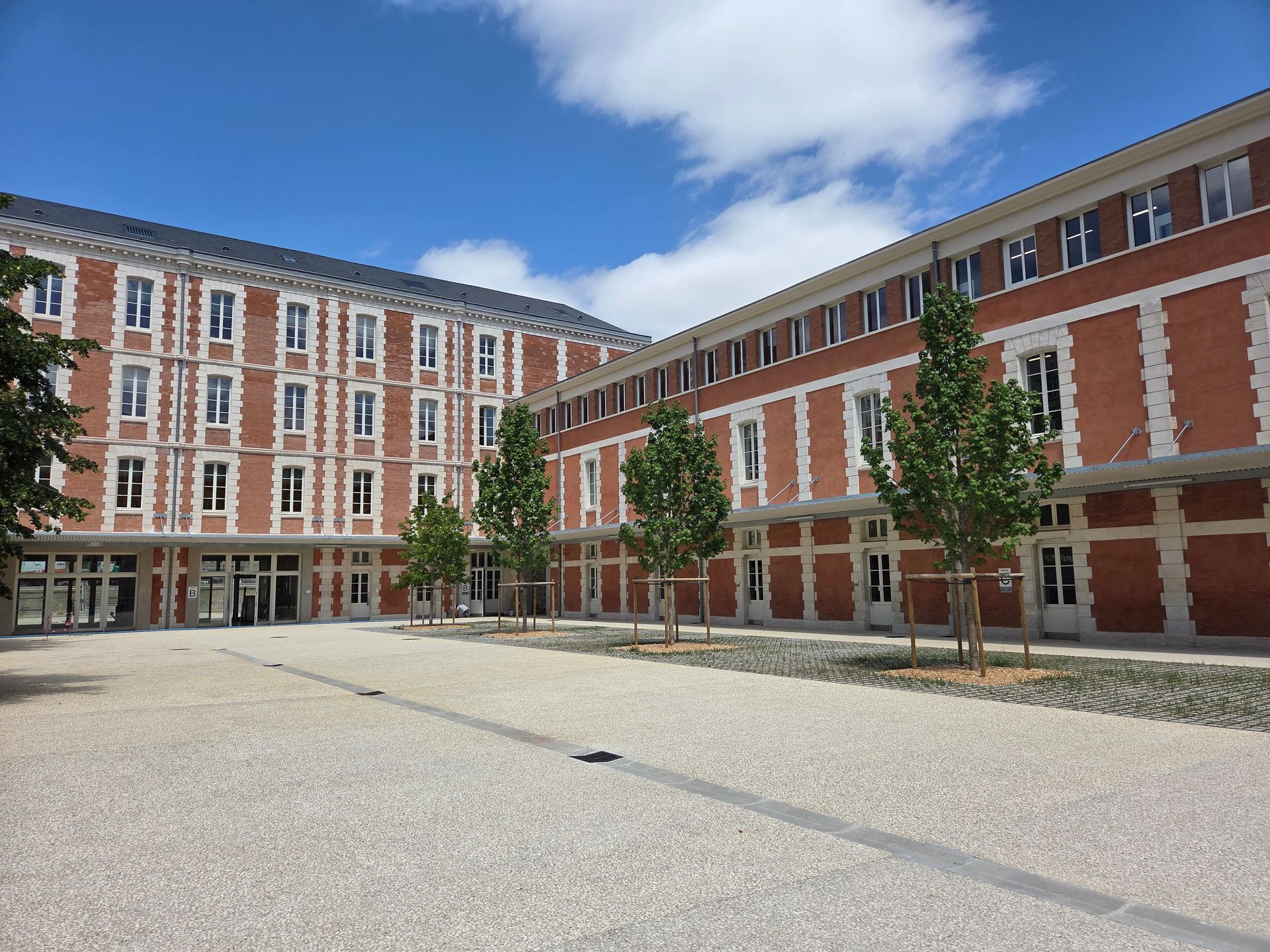 Cour intérieure moderne en briques, avec des arbres, un sol pavé et un ciel bleu entre deux bâtiments