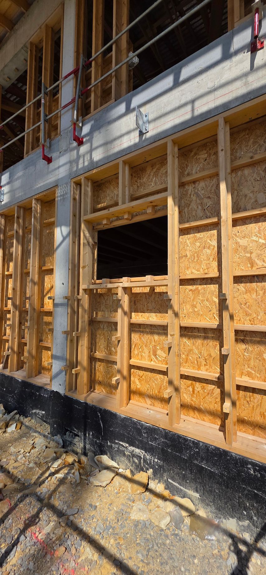 Ossature et revêtement extérieur d’un mur sur un chantier, avec une ouverture pour une fenêtre et un échafaudage au-dessus.