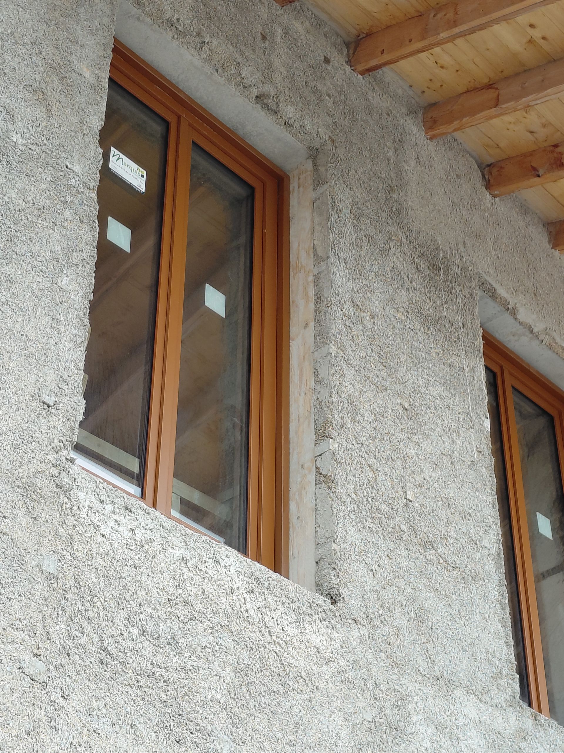 Fenêtres en bois encastrées dans un mur en crépi brut, sous des poutres apparentes au plafond