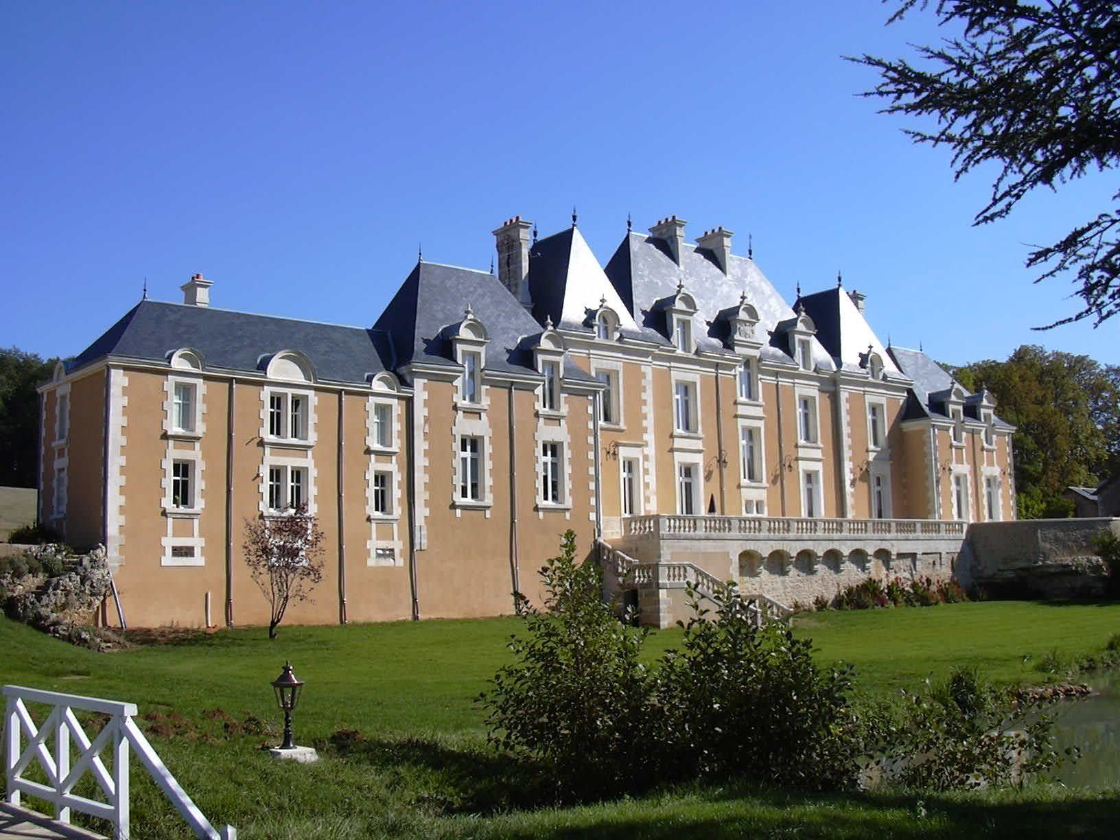 Grande demeure couleur sable, semblable à un château, avec des toits en ardoise grise, sur une pelouse verte sous un ciel bleu dégagé