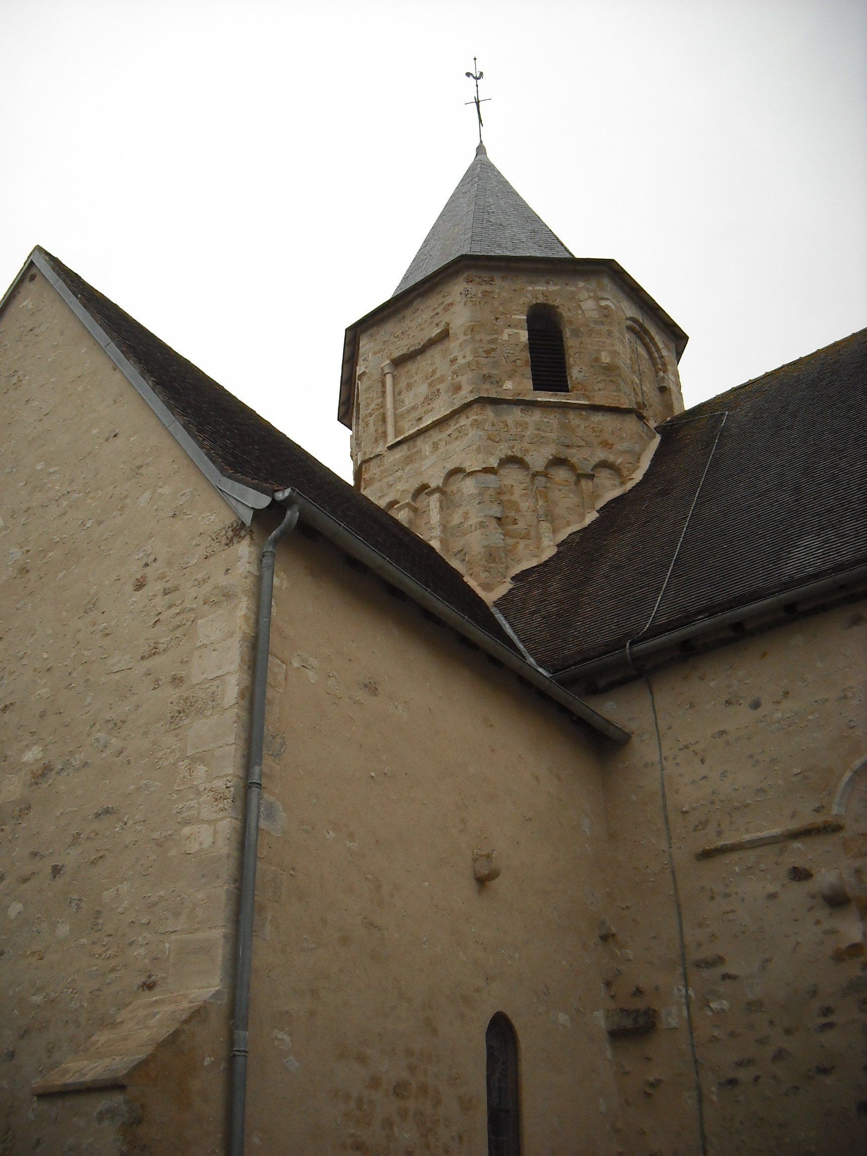Tour d’église en pierre surmontée d’un toit pointu, qui s’élève au-dessus de murs de bâtiment couleur sable et d’un toit couvert de tuiles sombres