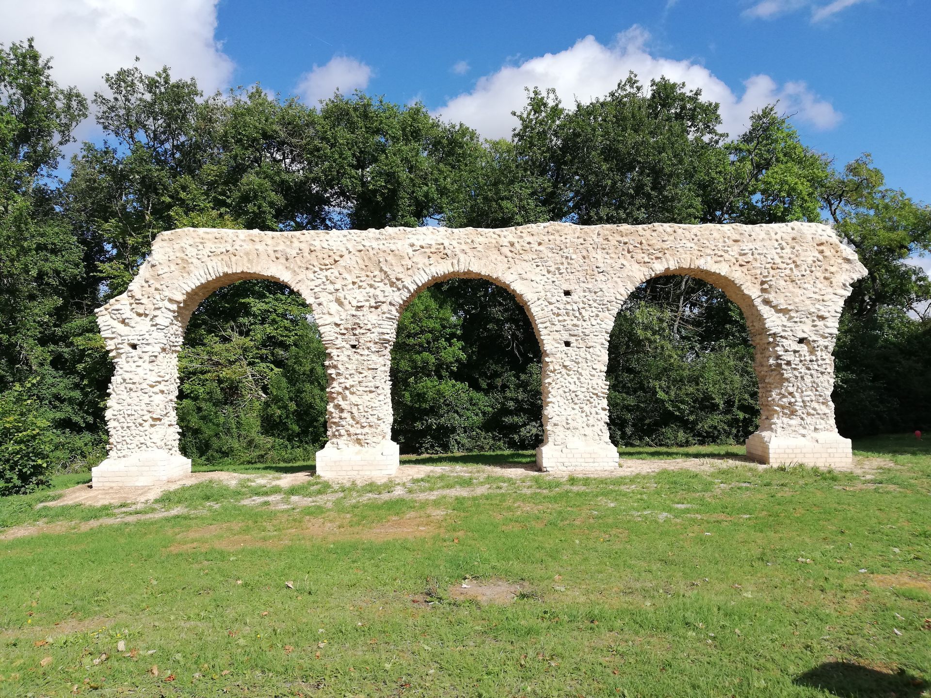 Ruines en pierre avec trois arches sur une colline verdoyante, sous un ciel bleu et avec des arbres en arrière-plan
