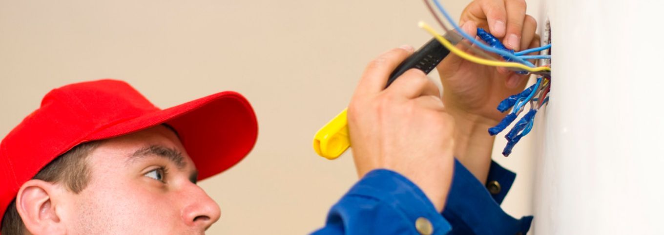 Electricista amb gorra vermella treballant amb cables, sostenint una eina groga a prop d'una paret blanca.