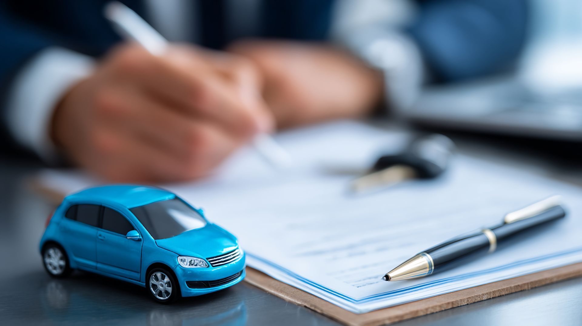 Une petite voiture bleue à côté d'un stylo sur un bloc-notes, avec une personne floue écrivant en arrière-plan.