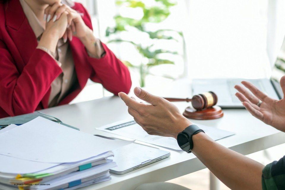 Iris Harff Rechtsanwältin, Frauenhand gestikuliert in der Luft über Schreibtisch mit Unterlagen gegenüber Oberkörper eine Frau mit am Tisch aufgestützten Händen