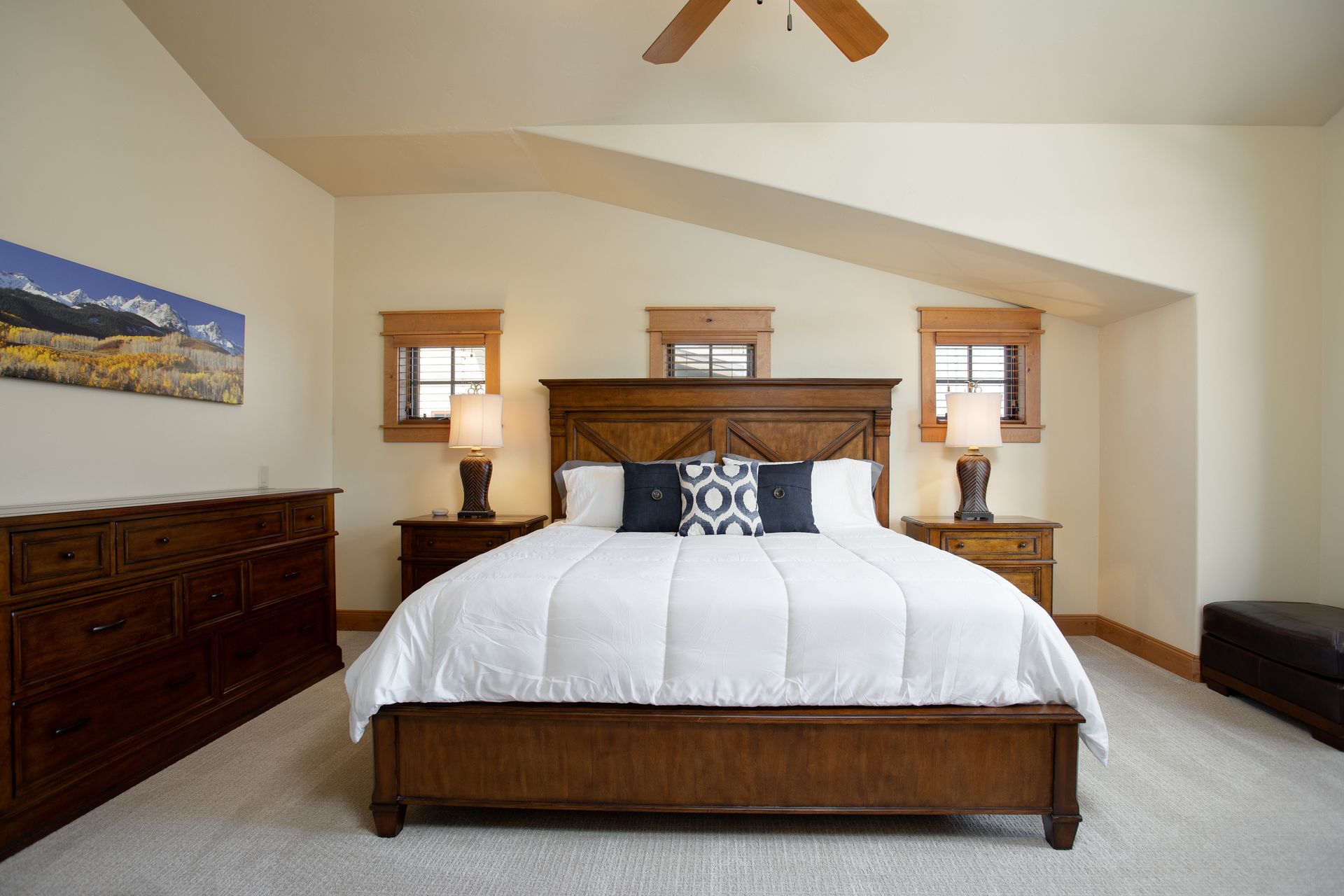Bedroom with a wooden bed, nightstands, windows, dresser, and a ceiling fan; beige walls, white bedding, and light carpet.