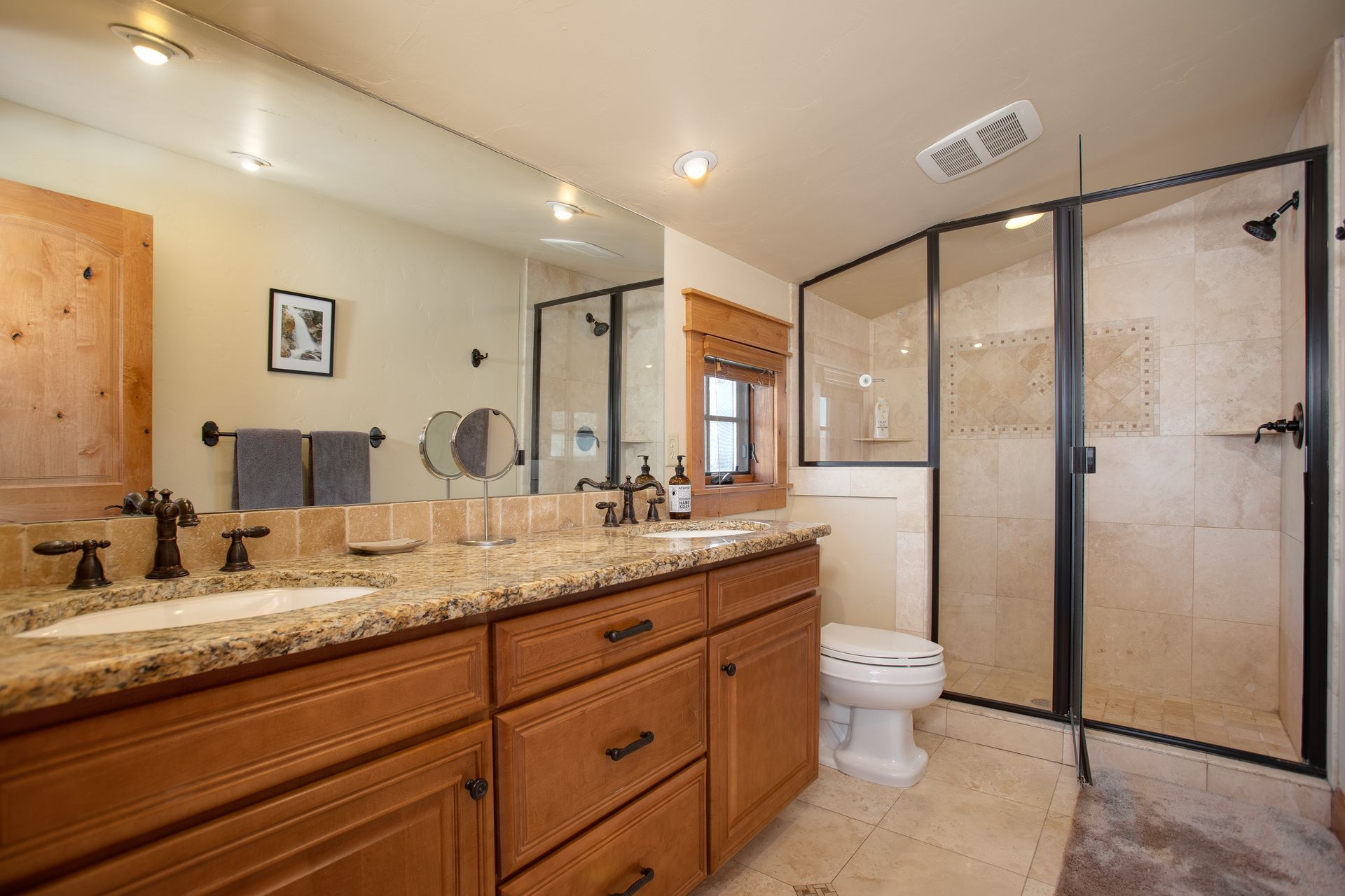 Bathroom with double sinks, large mirror, and glass-enclosed shower; light wood cabinets and beige tile.
