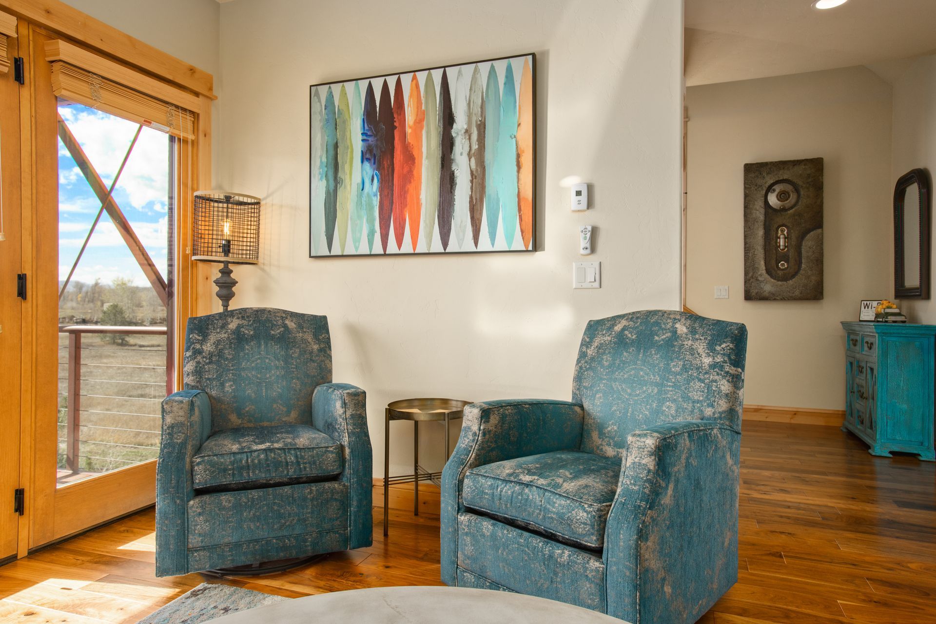 Two teal armchairs in a living room, artwork on the wall, wooden floors, and a view through a window.