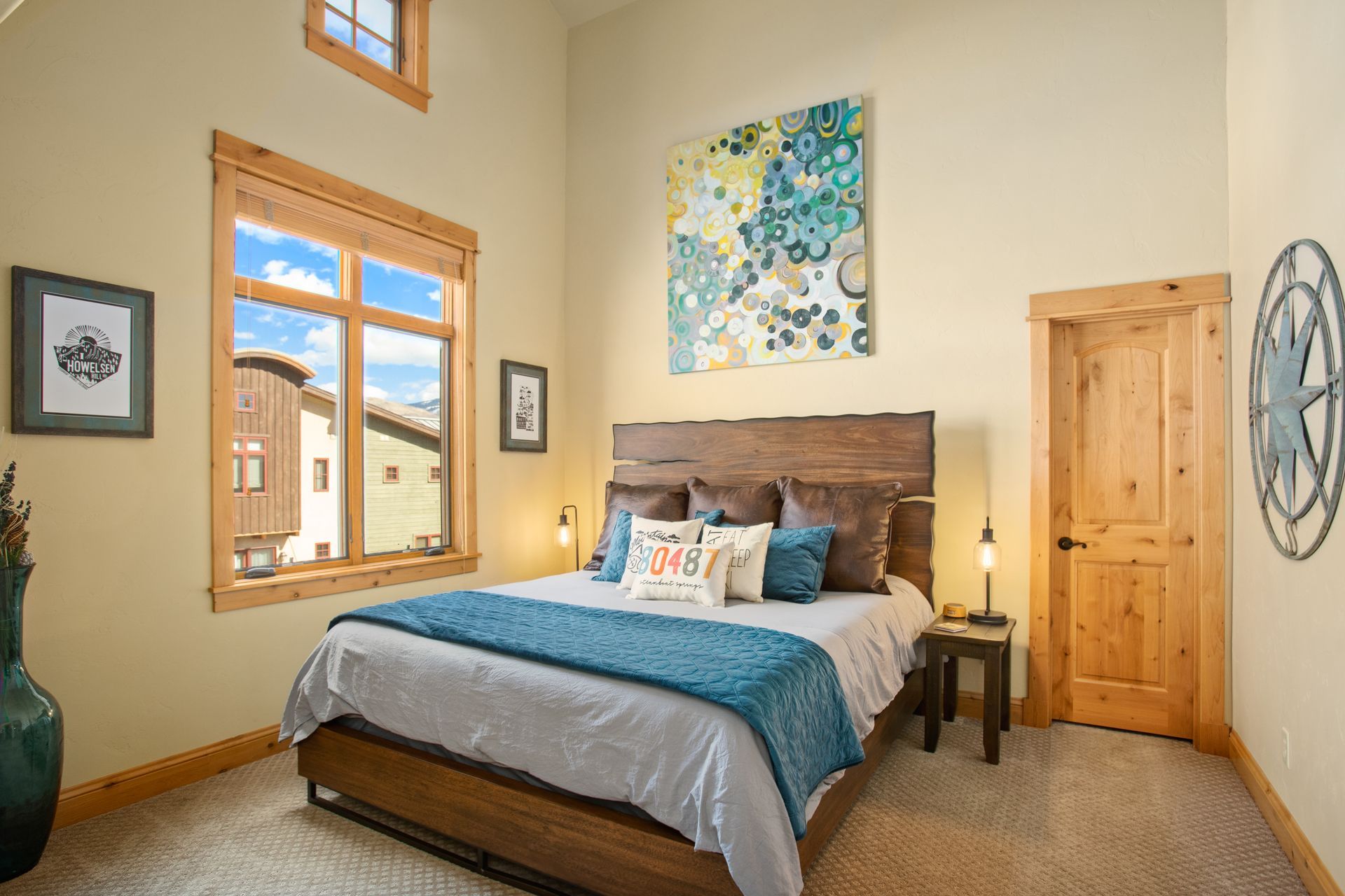Bedroom with bed, art, window, wooden door, and accents. Blue, brown, and cream colors.