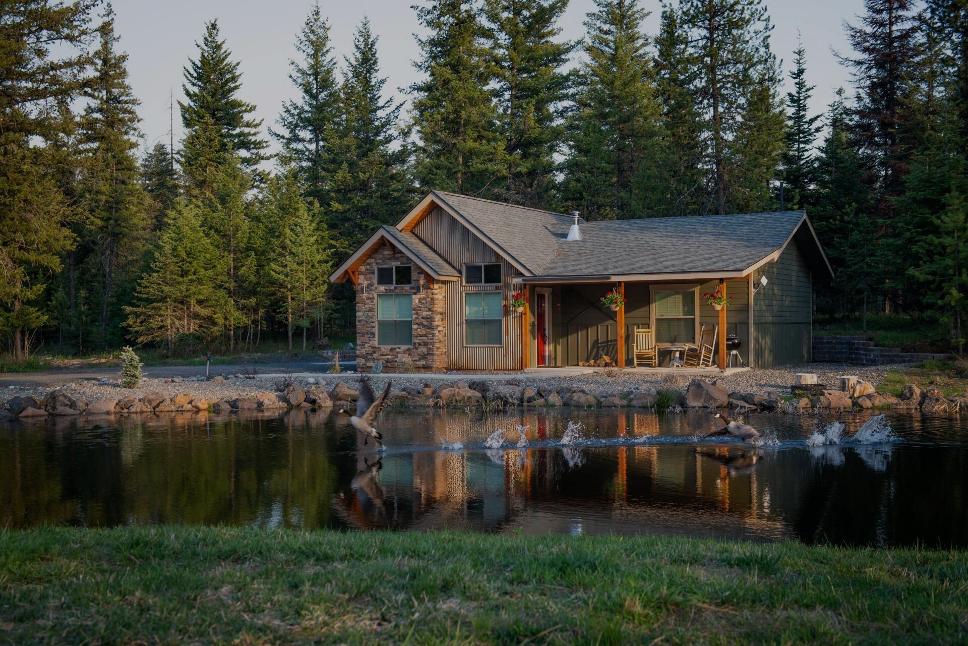 Cabin beside a calm lake, reflecting trees and the structure's stone and wood facade. Forest backdrop.