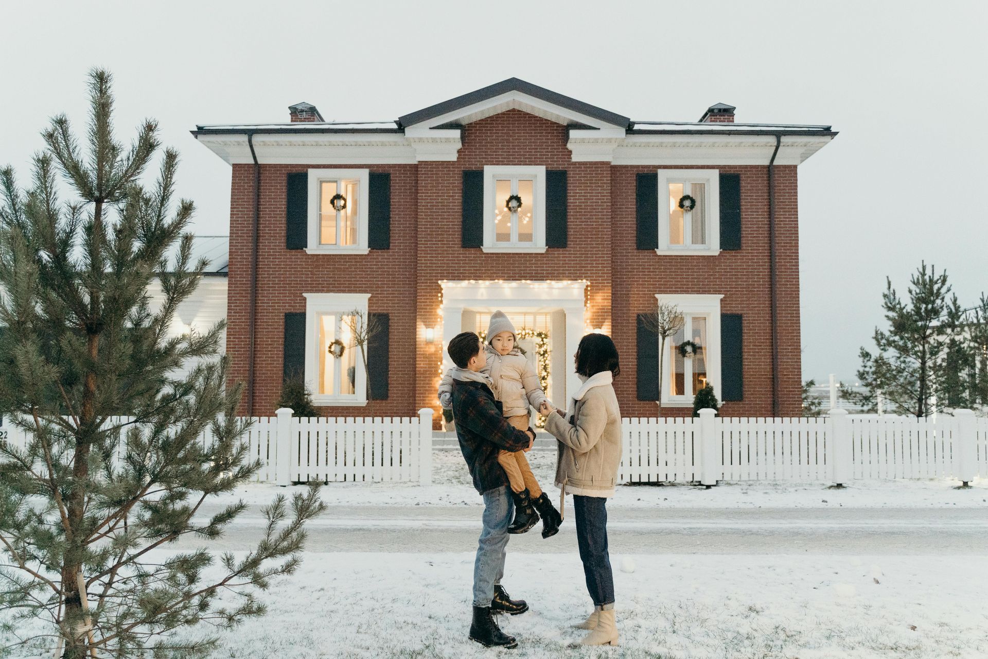 A family is standing in front of a large brick house in the snow.