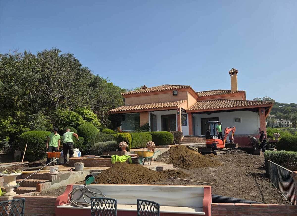 Obreros acondicionando el jardín delantero de una casa color beige con techo de tejas rojas.