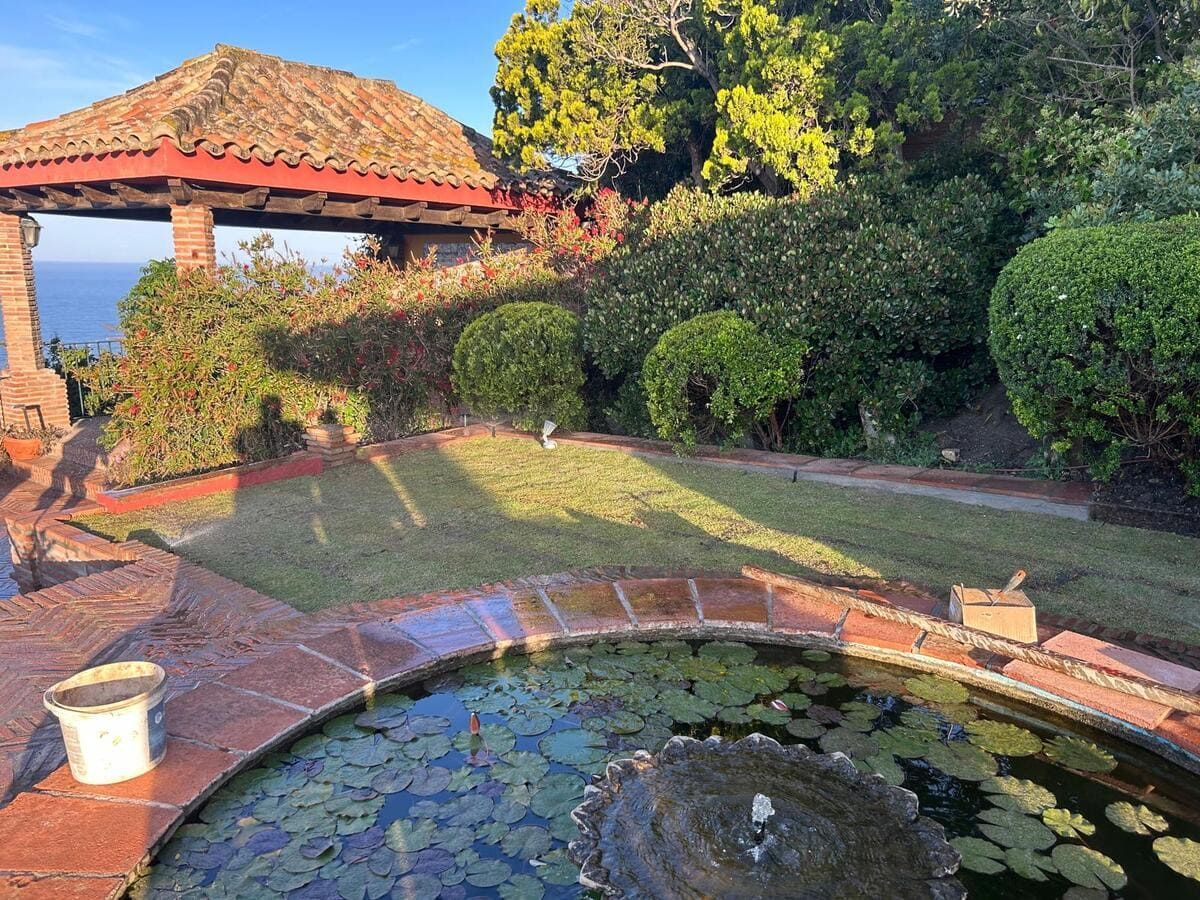 Estanque de jardín con nenúfares, borde de ladrillo y un cenador con tejado de tejas rojas junto a setos recortados.