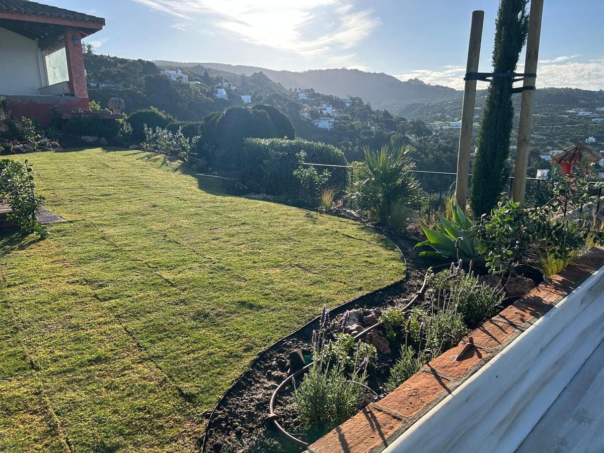 Jardín soleado en la ladera de una colina, con césped verde, borde de ladrillo y vistas al valle.